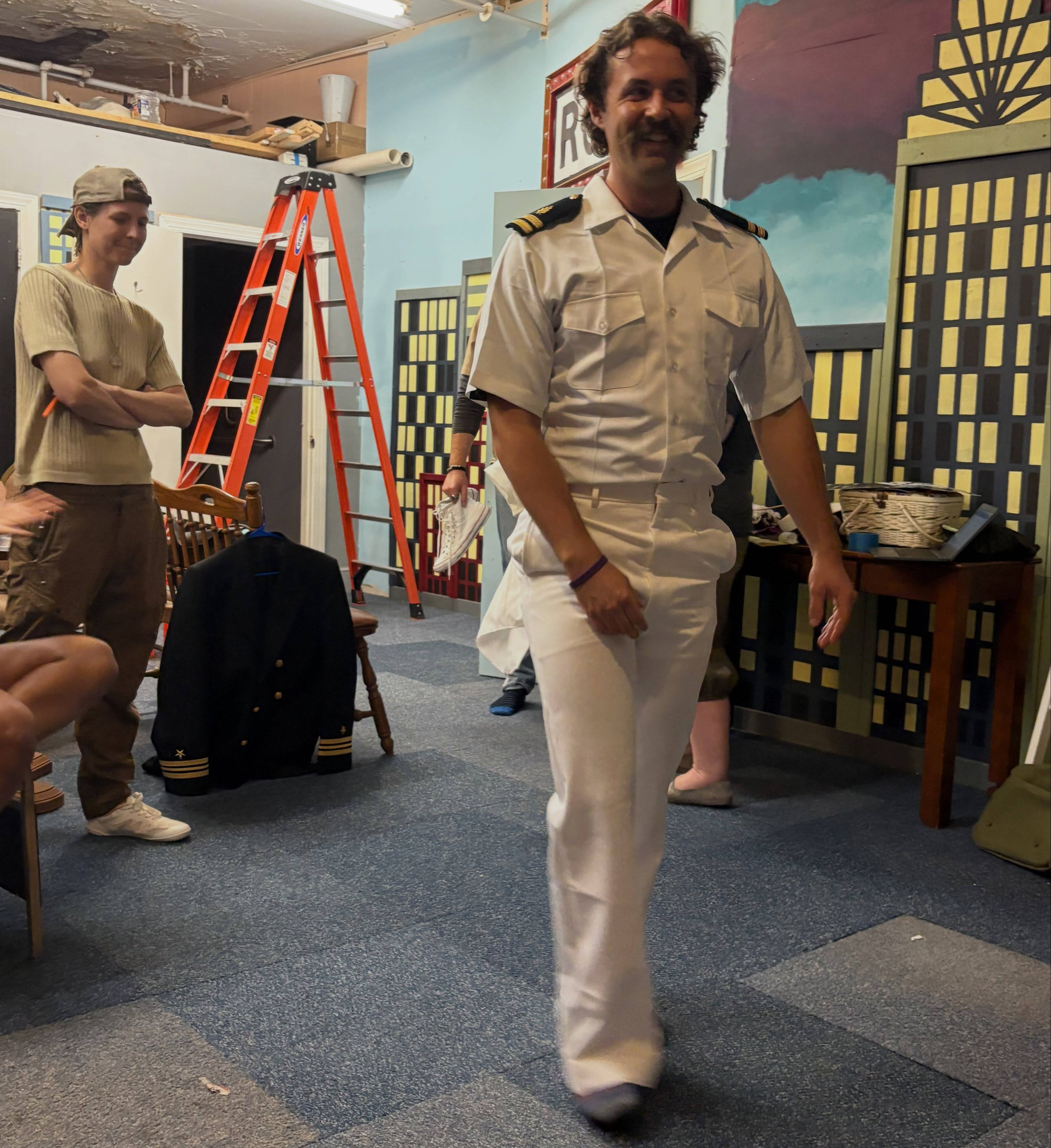 An actor tries on a white Navy officer’s uniform on the rehearsal room floor while a castmate looks on, costume racks and set flats around them.