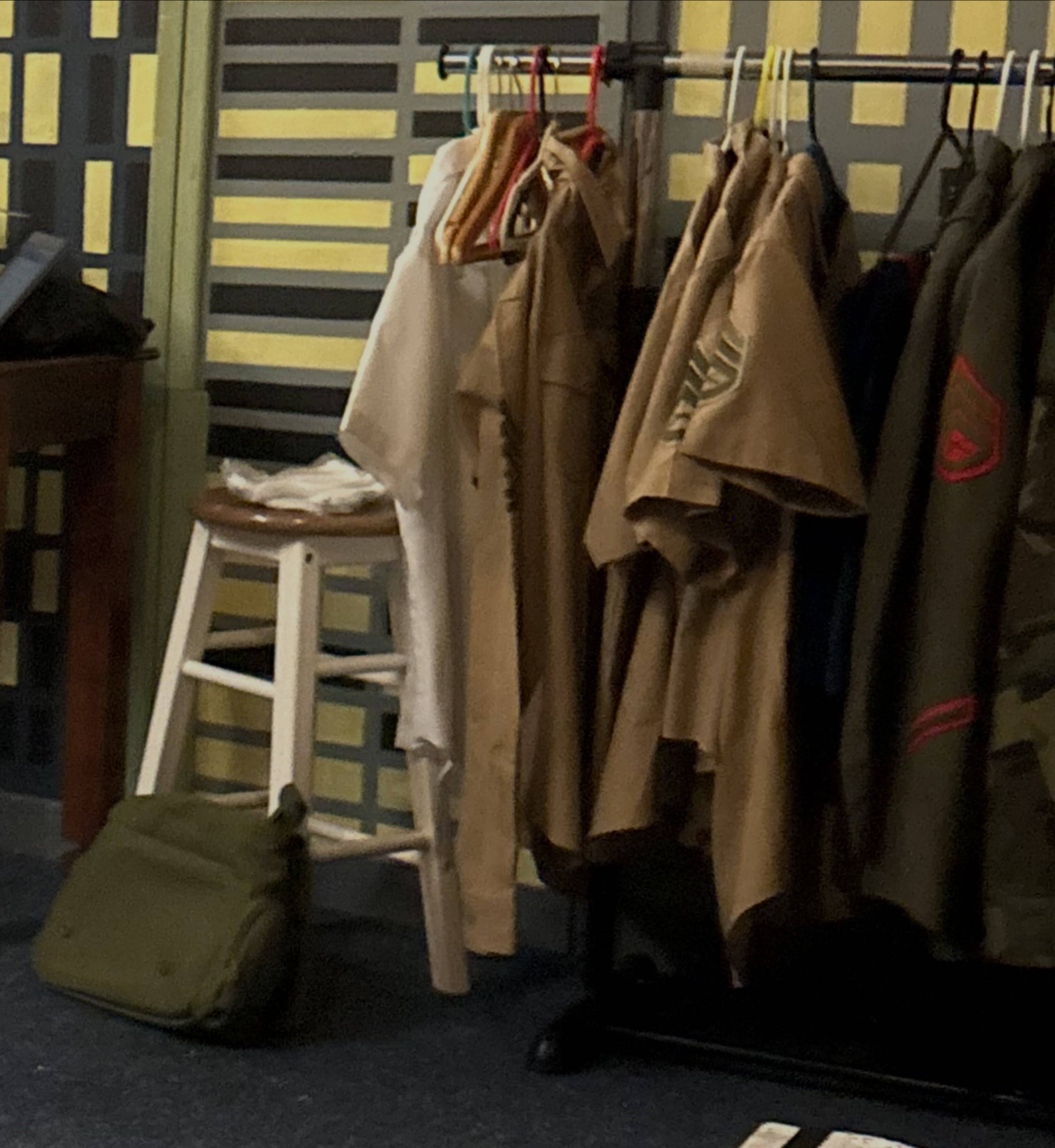 Khaki and camouflage military uniforms on a rolling costume rack backstage, a duffel bag on the floor beside a stool.