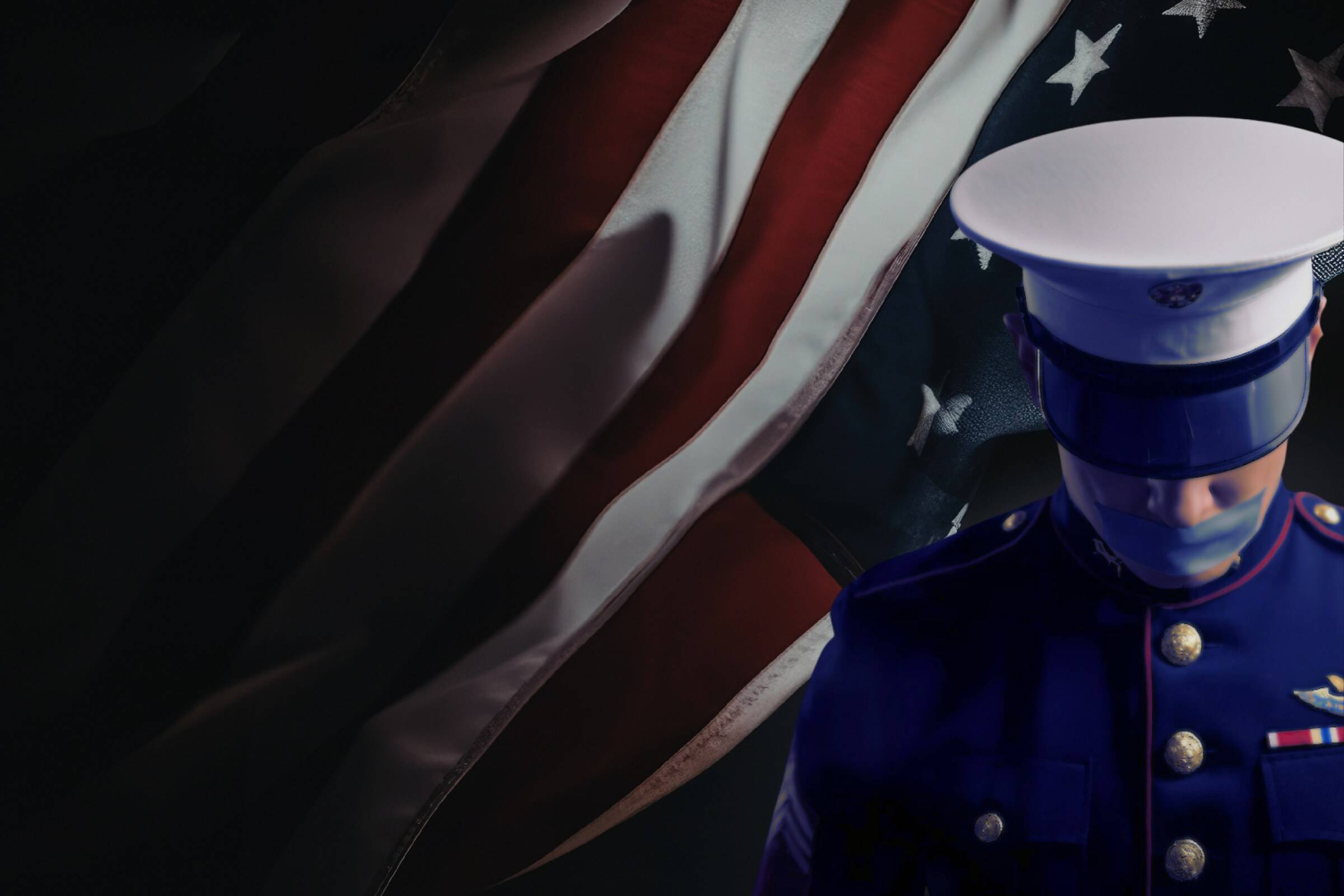 A Marine in dress blues and a white peaked cap, head lowered, standing in front of a hanging American flag in deep shadow