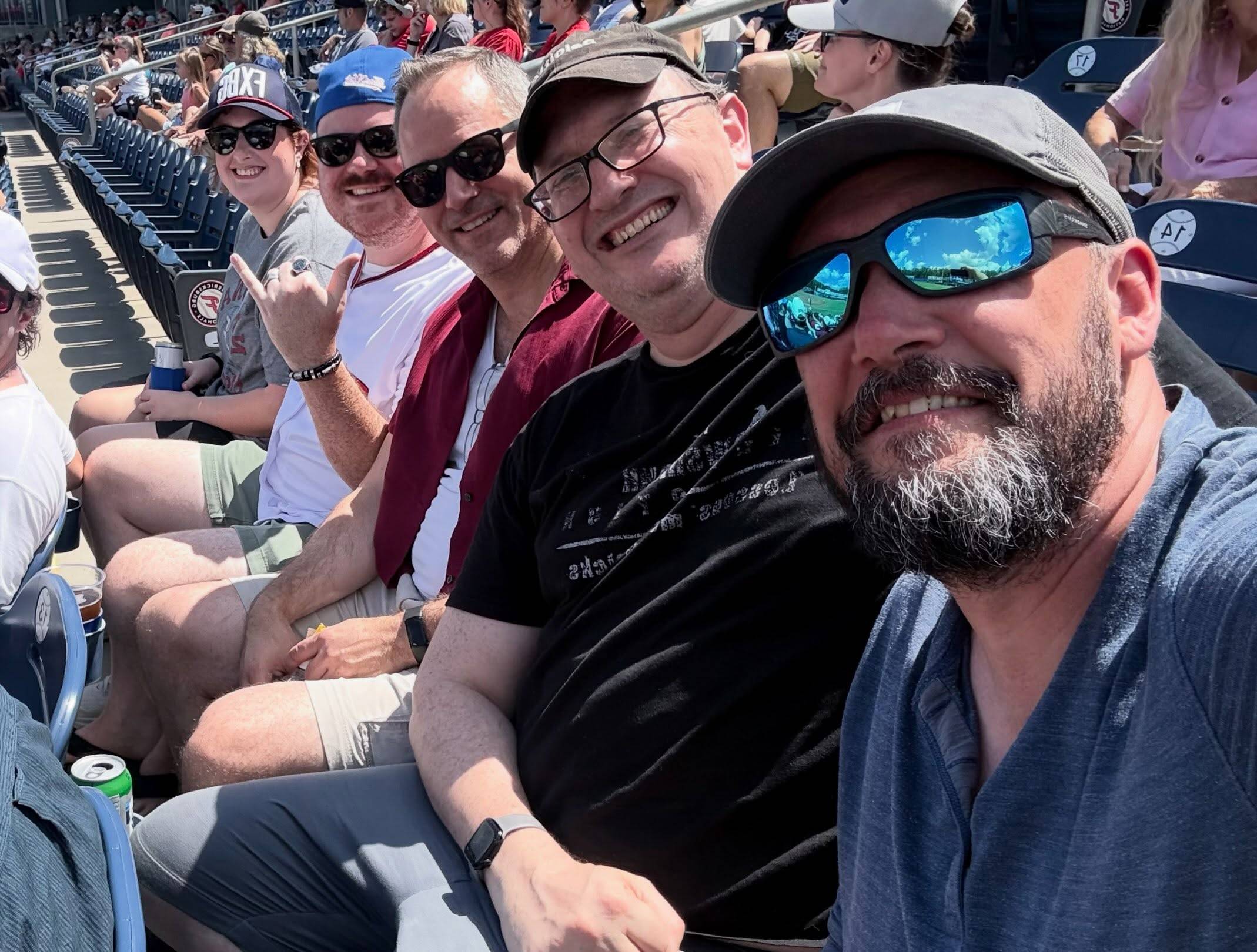Five members of the A Few Good Men cast and crew squeeze into a selfie in the stadium stands, all smiles in caps and sunglasses.