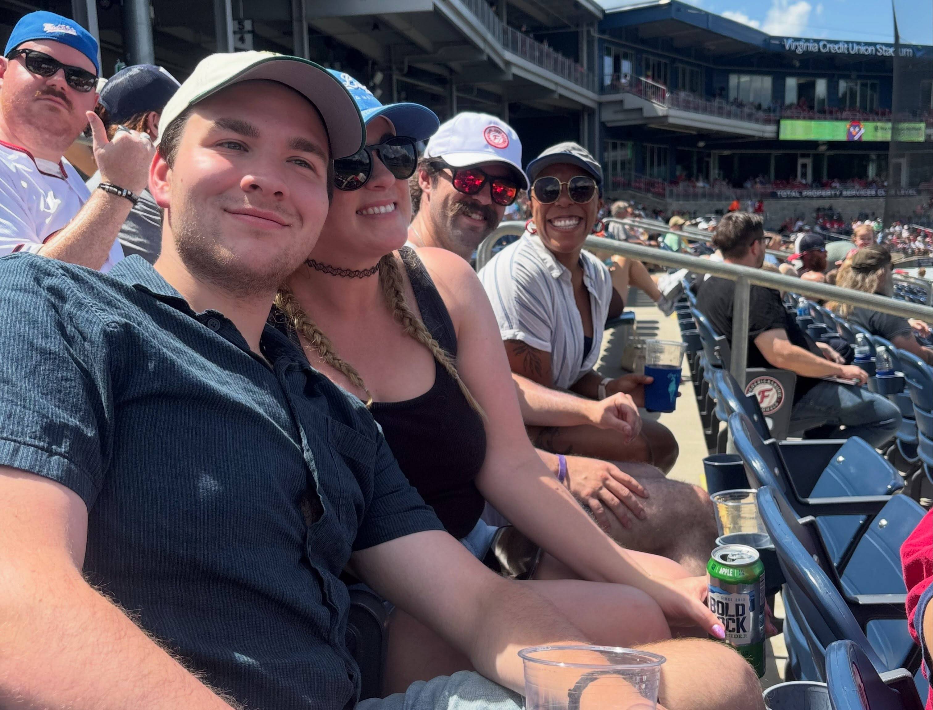 Four cast members sit along a row of ballpark seats on a sunny afternoon, drinks in hand.