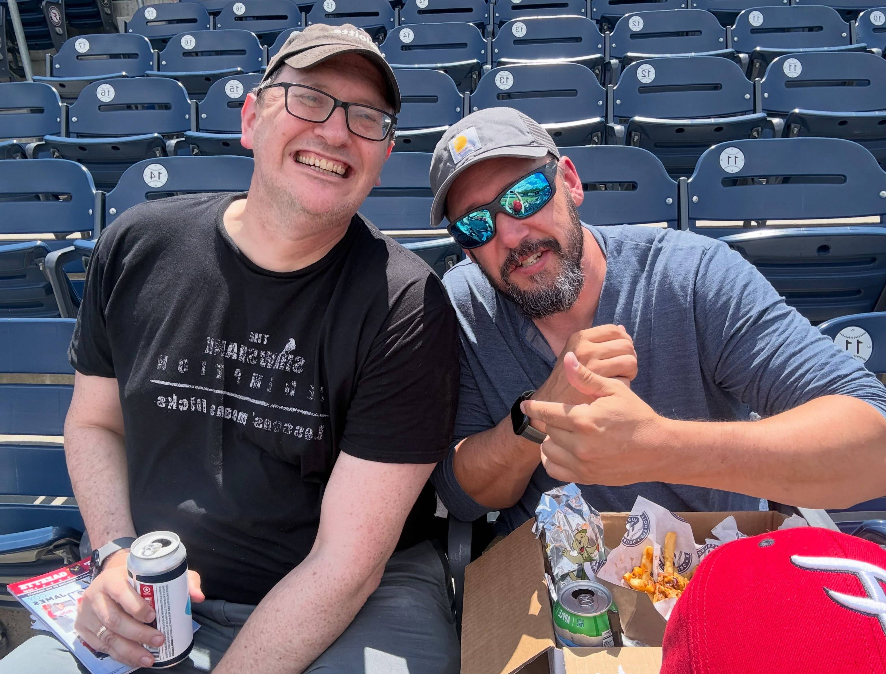Two cast members lean in for a photo in the stands with a tray of ballpark fries and drinks in front of them.