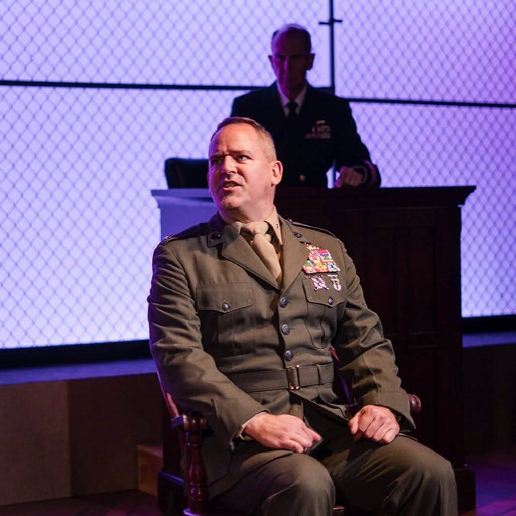 A performer in a Marine dress green service uniform with ribbon bars and badges sits in a wooden chair on the witness stand, head turned, mouth open mid-line. Behind and above them another performer in a dark naval service dress coat with ribbons sits at a raised wooden bench. A chain-link wall lit lavender fills the back of the stage.