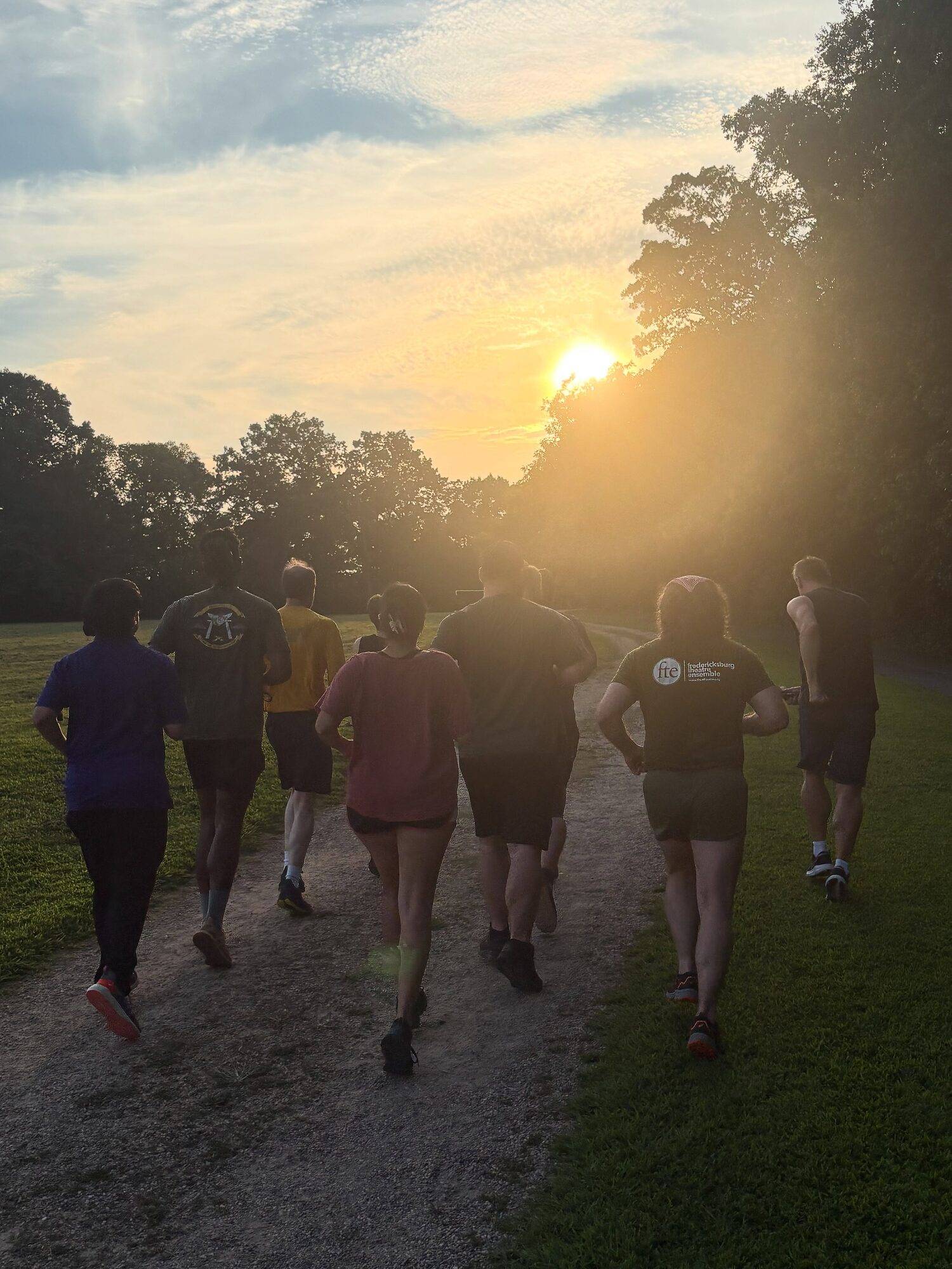 Cast and crew jog down a gravel park path toward the setting sun, one runner in a Fredericksburg Theatre Ensemble shirt.
