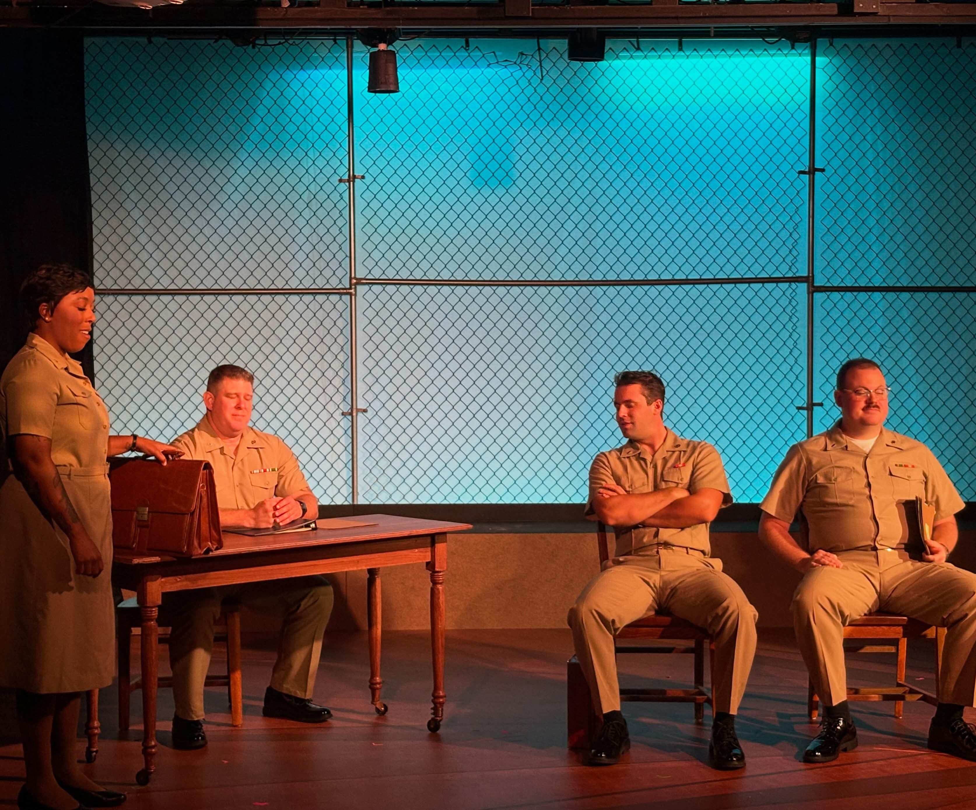 Four performers in tan service uniforms in a courtroom scene lit turquoise. One stands at the left beside a wooden table with a leather briefcase on it, speaking; another sits at the table with hands folded; two more sit on chairs at the right, one with arms crossed, the other holding a folder.