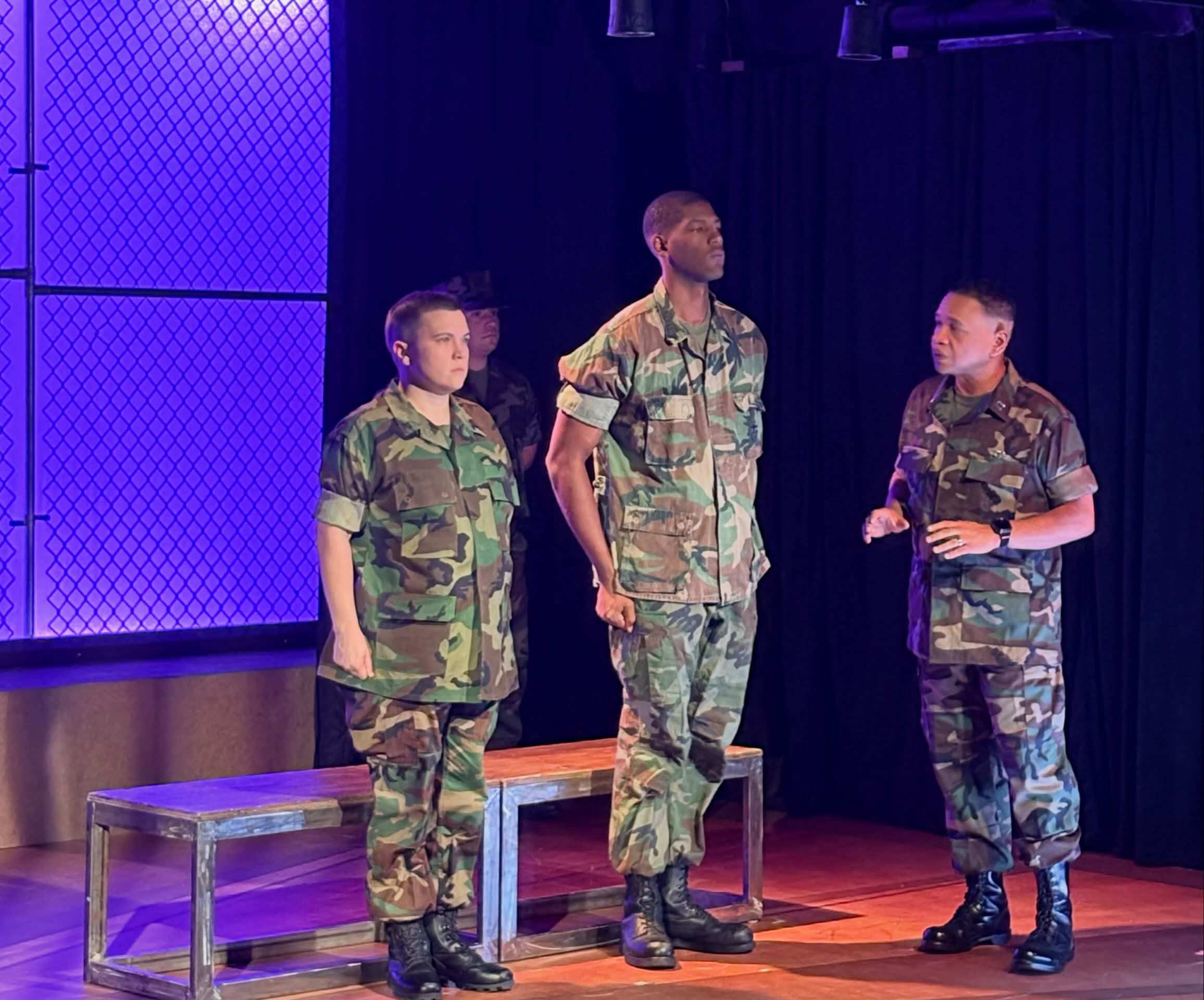 Three performers in camouflage utilities stand on a wooden stage lit purple and orange, two at attention in front of low benches while the third, at the right, turns toward them speaking with one hand raised. A fourth performer stands in shadow upstage.