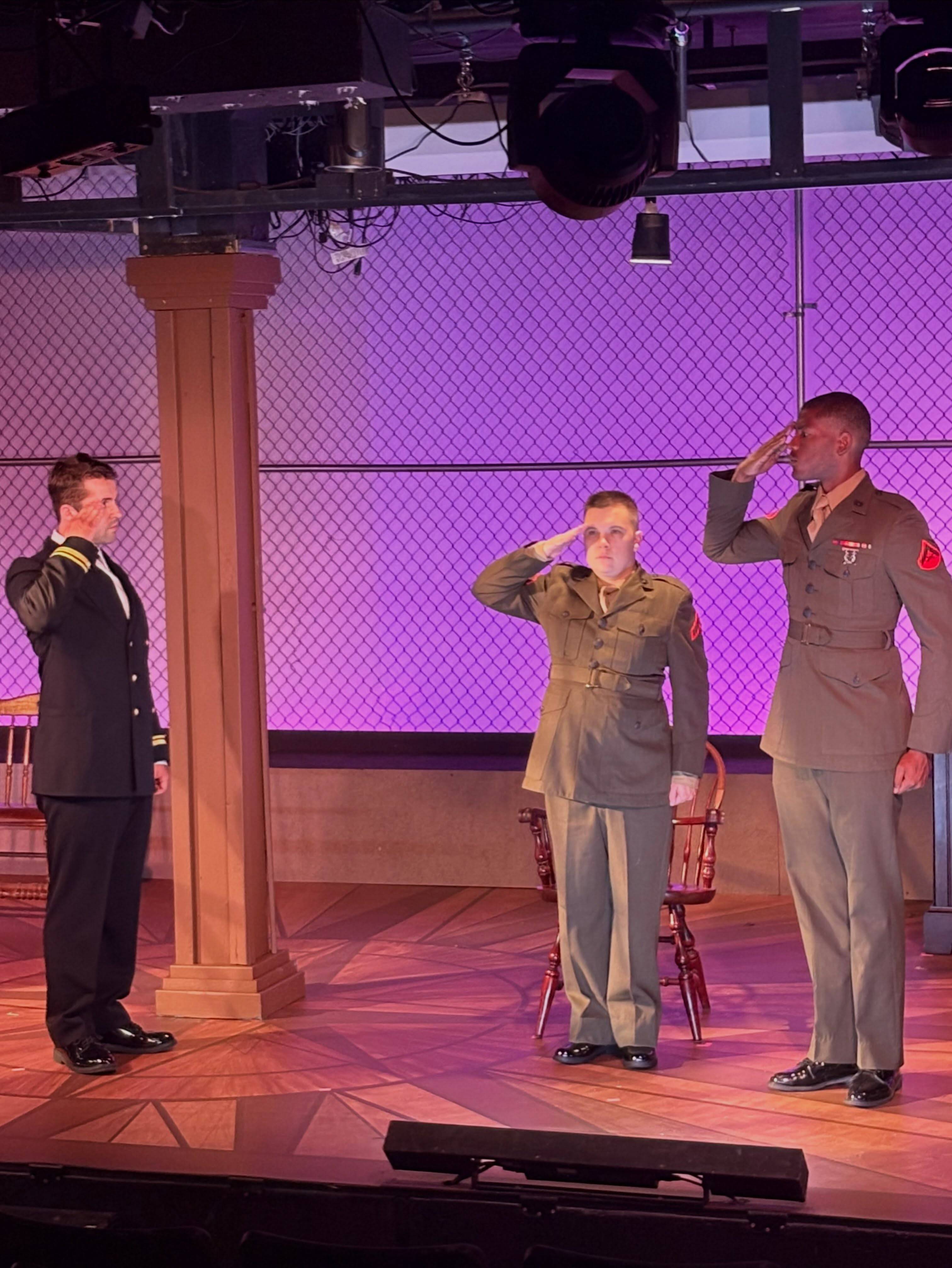 Three performers salute on stage before a purple-lit chain-link wall, one in Navy dress blues on the left and two in green Marine uniforms on the right, a wooden column and a chair between them.