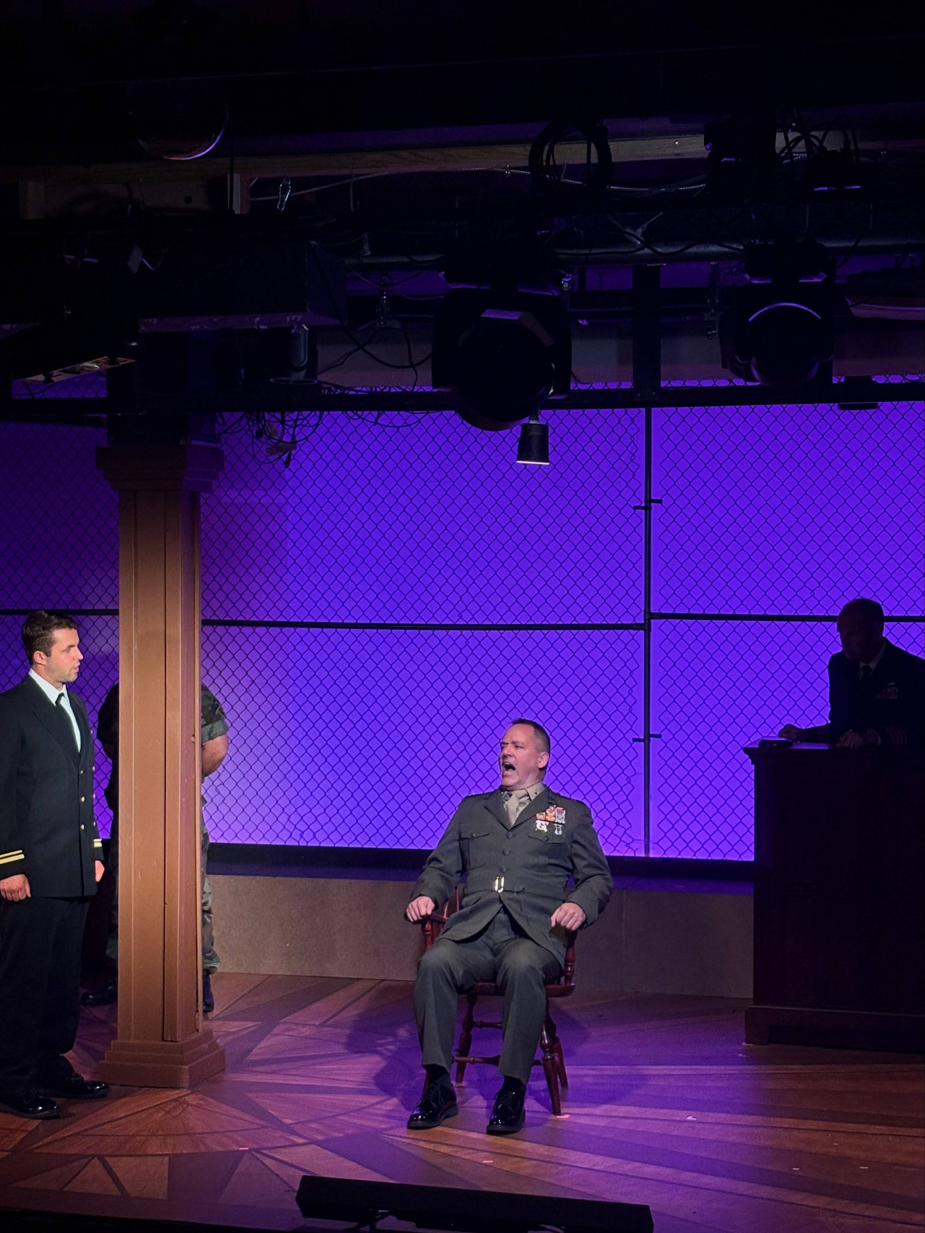 A performer in a green Marine dress uniform with medals sits in a chair centre stage, mouth open mid-line, under purple light. A performer in Navy dress blues stands at the left edge and another sits behind a raised bench on the right.