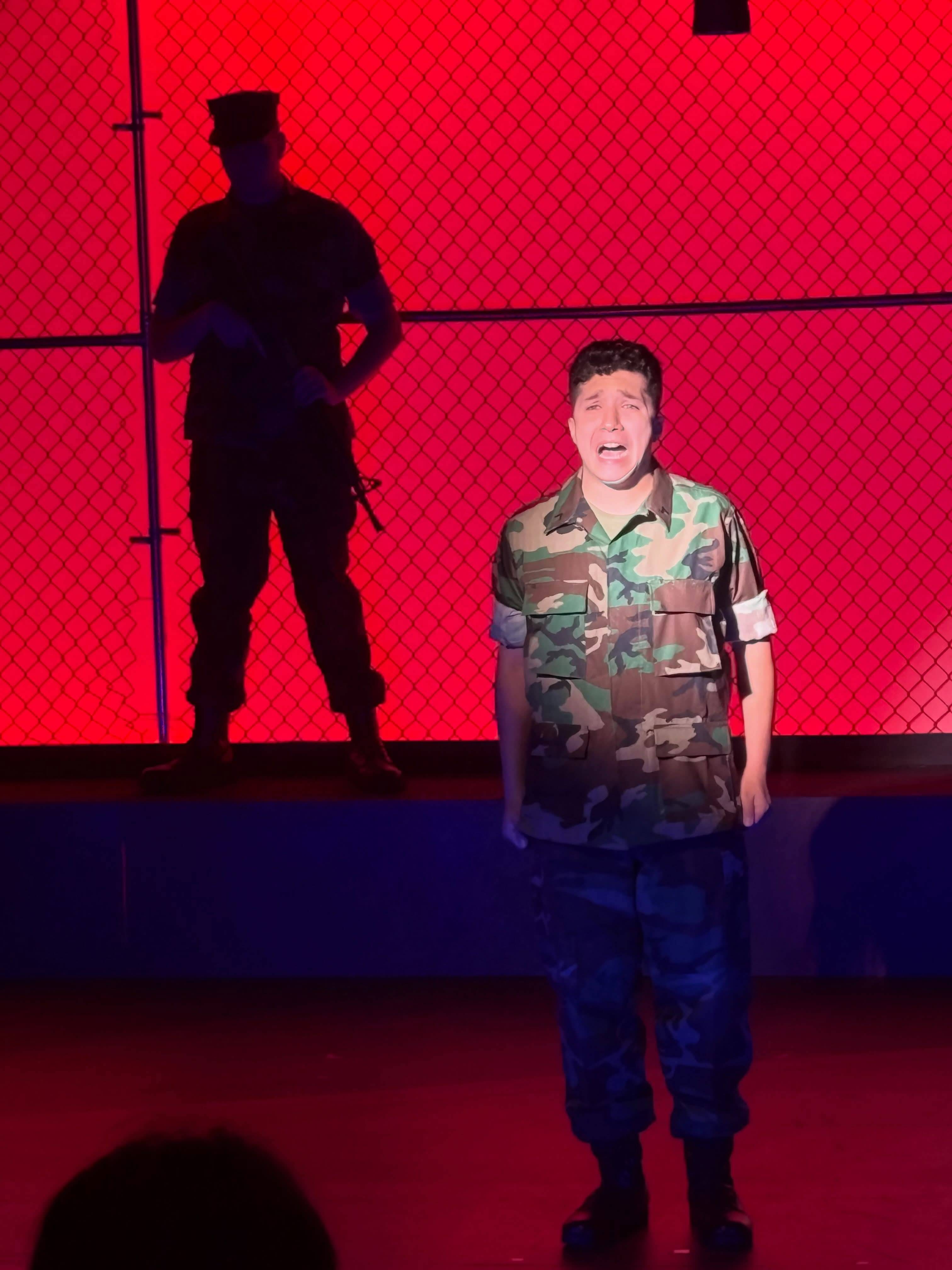 A performer in camouflage utilities stands centre stage mid-line, mouth open, in front of a chain-link wall lit bright red. Upstage on a platform another performer stands in silhouette in cover, holding a rifle across their body.