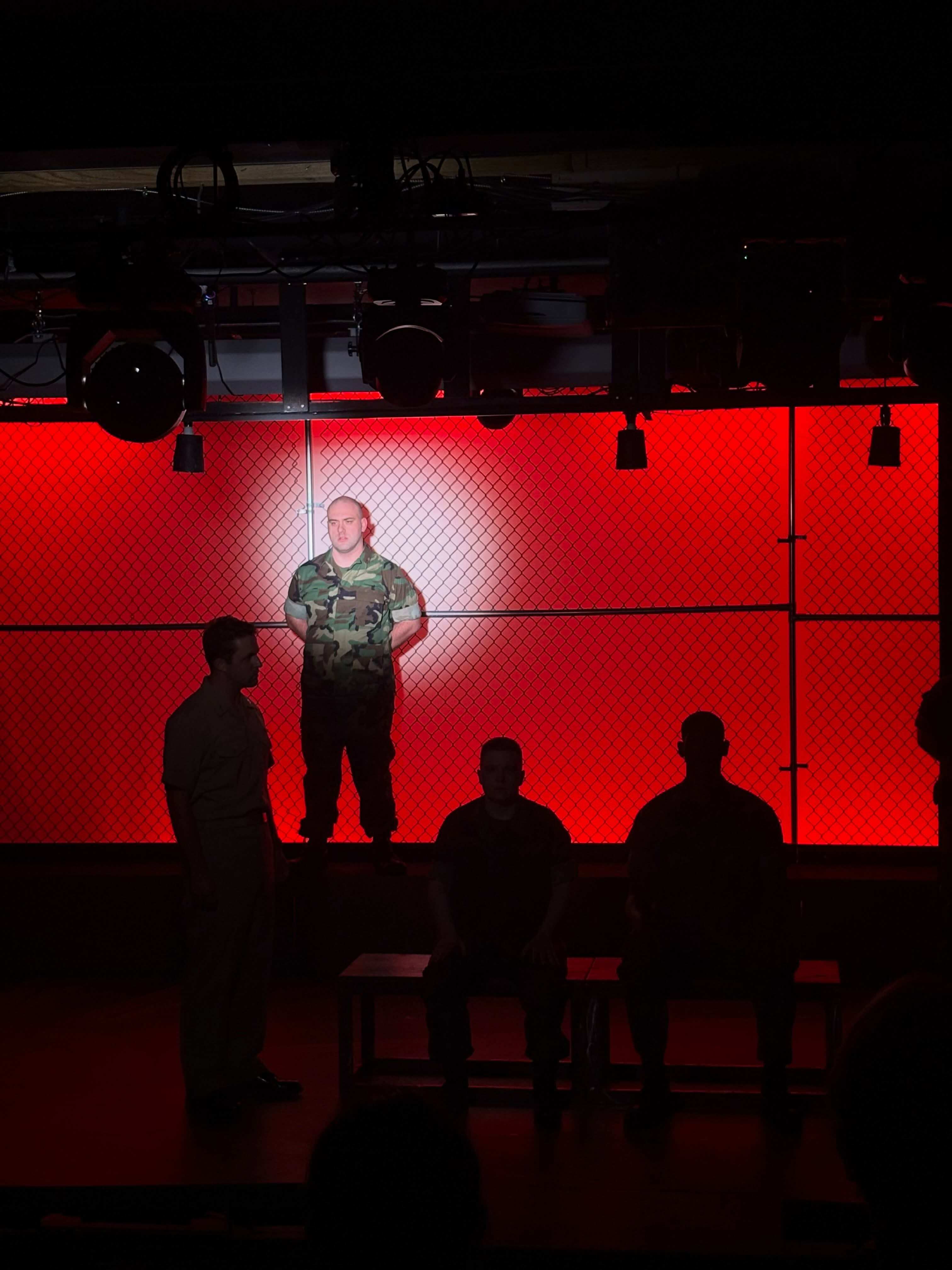 A performer in camouflage utilities stands at attention on a platform in a pool of white light, against a chain-link wall lit bright red. Three performers are in silhouette below: one standing at the left, two seated on benches. The lighting rig hangs across the top of the frame.