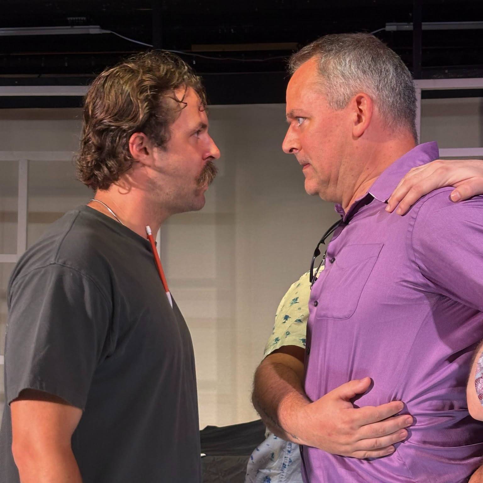 Two performers face off nose to nose on a black-box stage: one in a grey T-shirt and thin chain, the other in a purple shirt with sunglasses hung at the collar, a third person part-hidden between them.