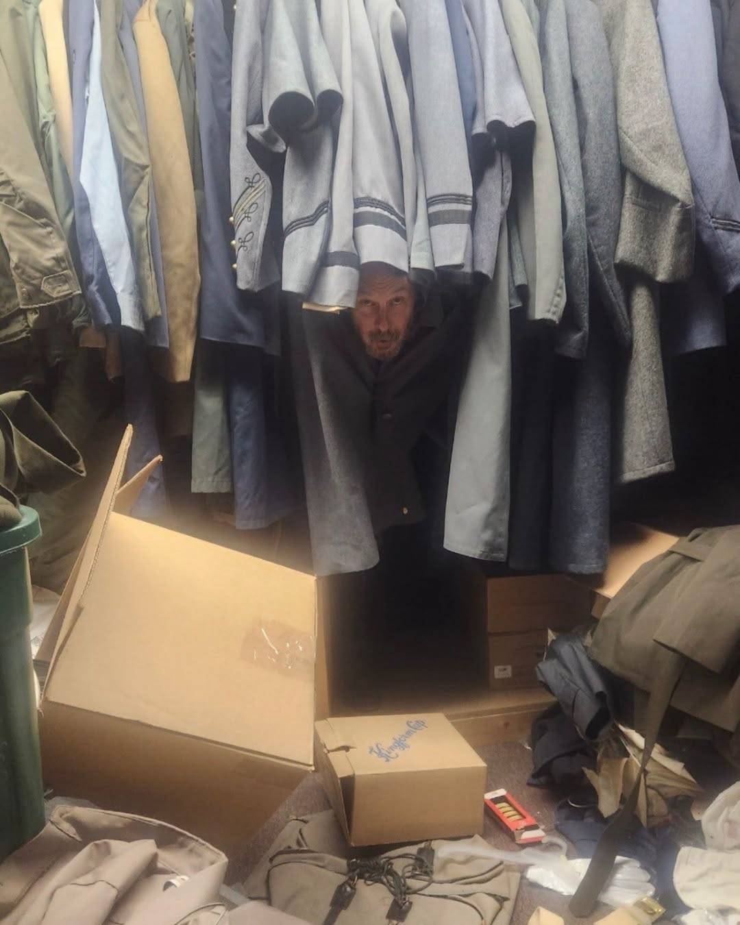 A member of the company peers out from between two packed rails of military uniforms, surrounded by open cardboard boxes.