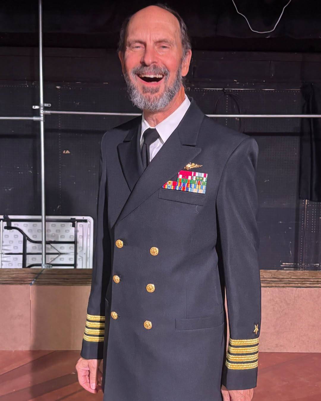 A performer laughs on stage in a Navy dress blue jacket with gold sleeve stripes, ribbon bars and wings on the chest.