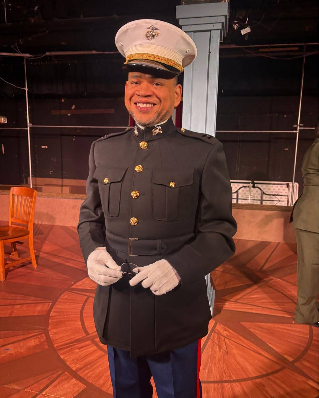 A performer smiles on stage in Marine dress blues with a white cover, white gloves and glasses held in hand.