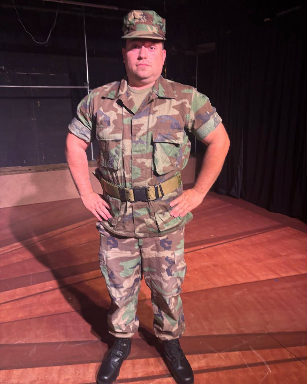 A performer stands at ease on stage in woodland camouflage utilities, sleeves rolled, with a cap and boots.
