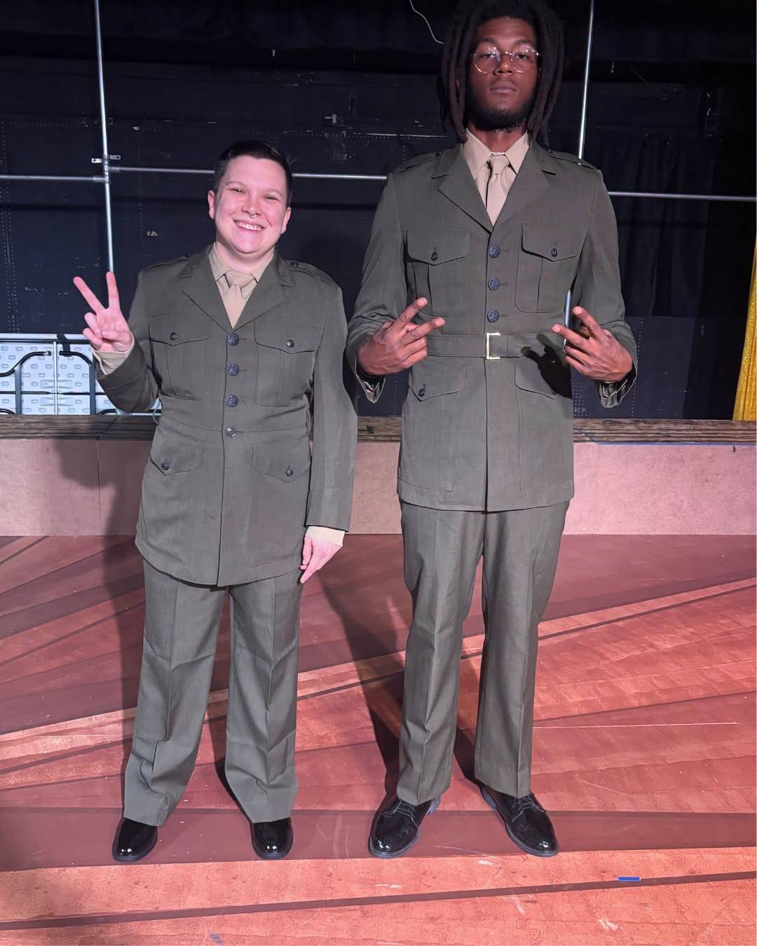 Two performers in olive green Marine service uniforms throw peace signs on stage.