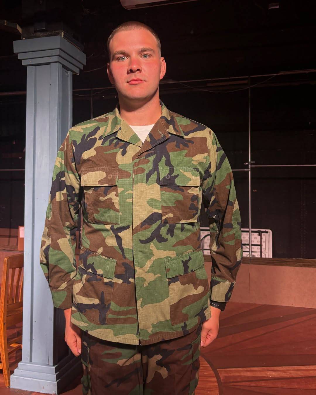 A performer stands on a bare stage in woodland camouflage utilities with the sleeves down, a white undershirt at the collar, beside a grey structural post.