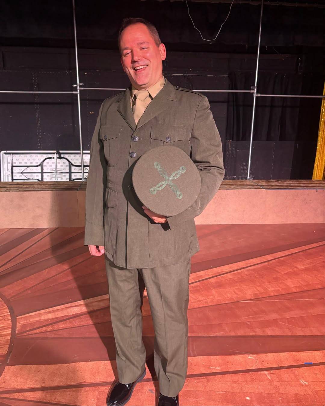 A performer laughs on stage in an olive green Marine service uniform with a khaki shirt and tie, holding a garrison cap with a crossed-braid device against their chest.