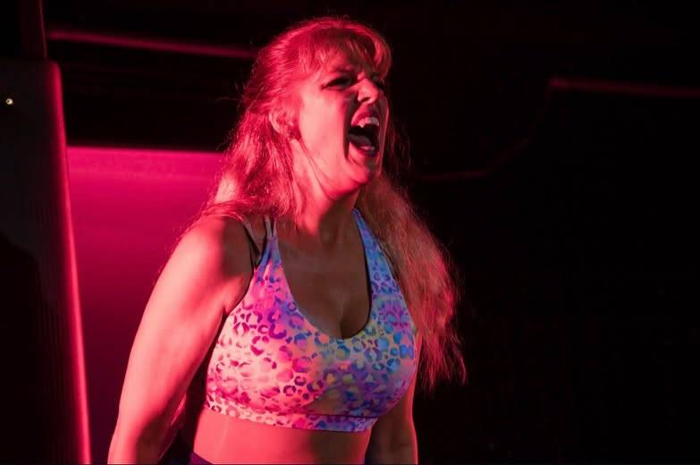 A dancer in a bright patterned sports bra roars with head thrown back under pink and red light.