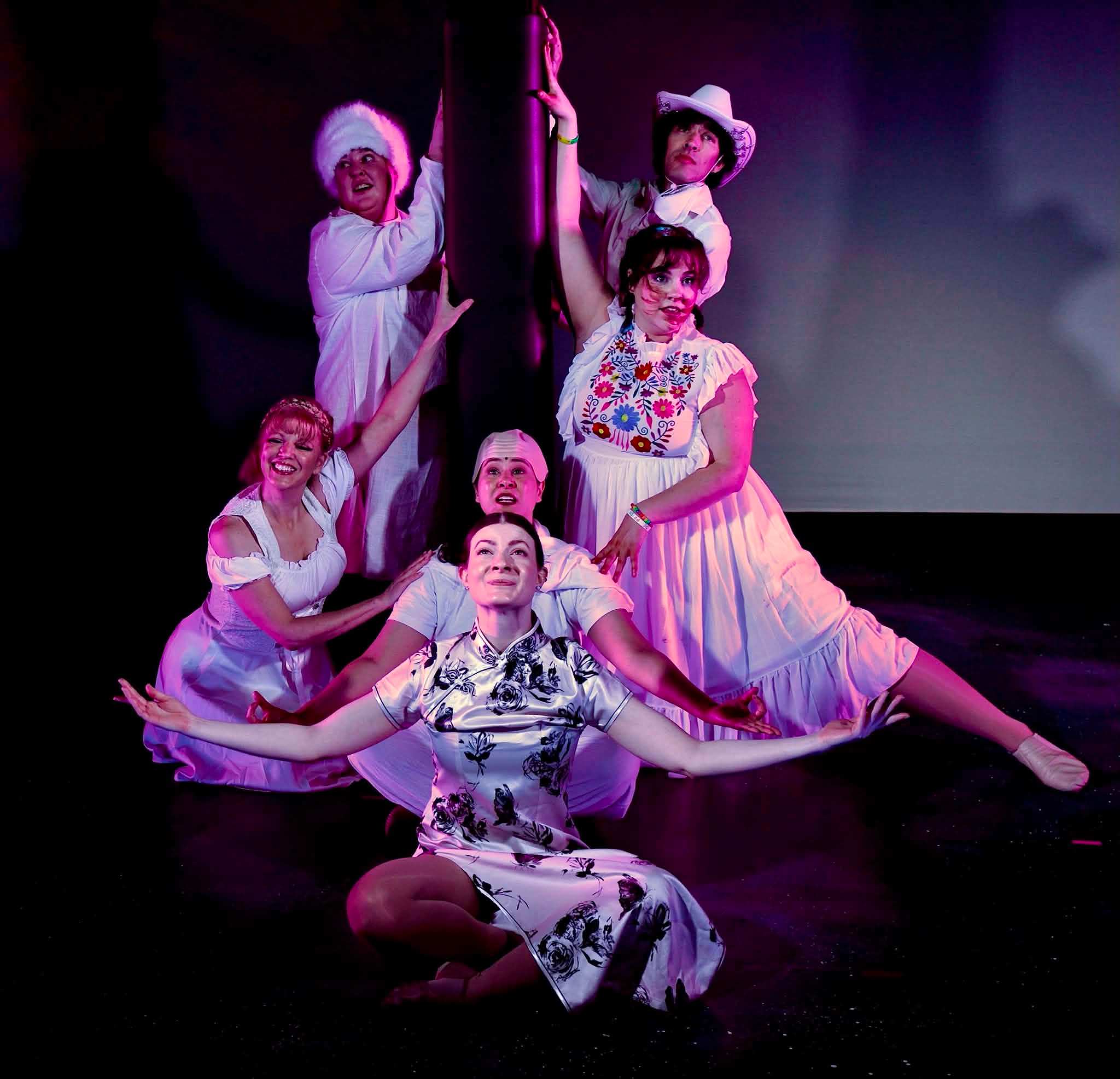 Six Dance Nation performers hold a theatrical group pose in white costumes under pink stage lighting.