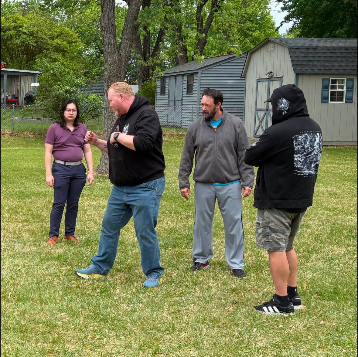 Four people stand on a lawn working through a stage fight, one stepping forward with a fist raised while the others watch. Garden sheds and a chain-link fence are behind.