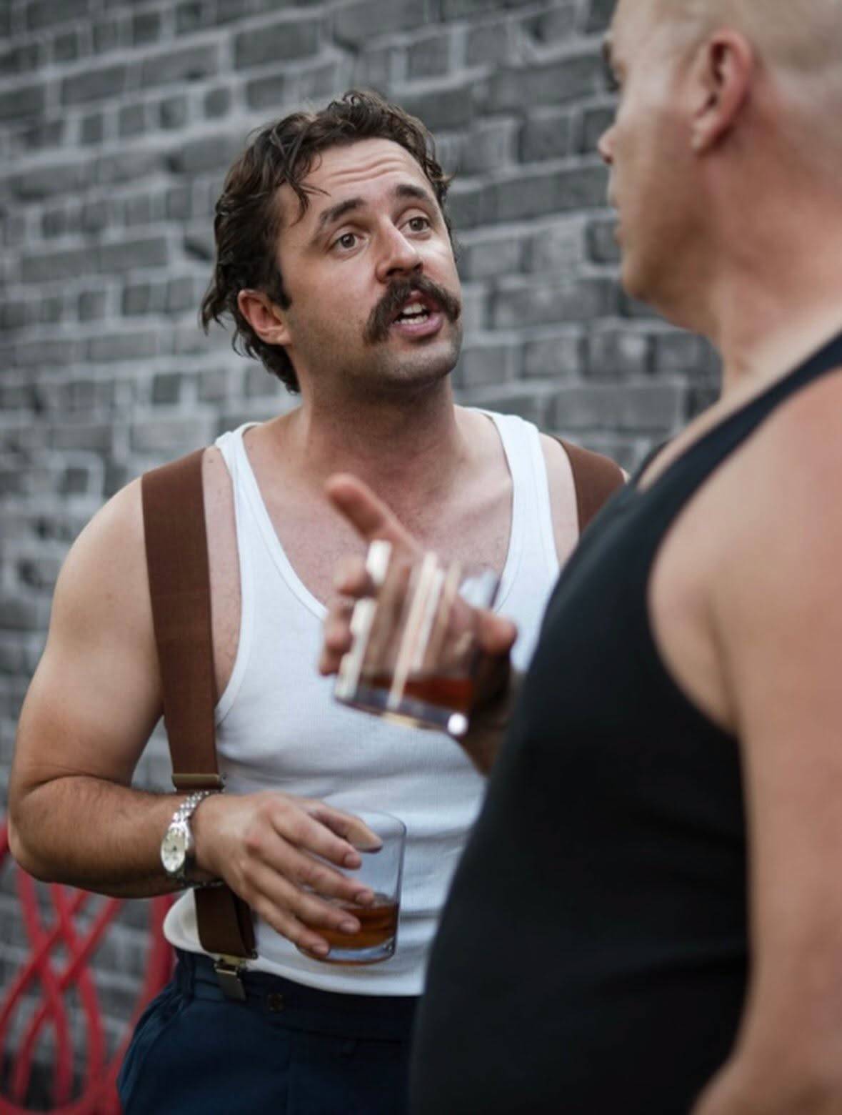 A performer in a white vest and brown braces, a watch on one wrist, holds a tumbler of whisky and gestures mid-line toward another performer in a black vest seen from behind in the foreground. A grey brick wall is behind.