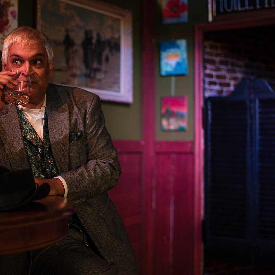 The performer sits alone at a small round table drinking from a glass, a black hat on the table in front of them, looking off to one side. The swing door and the pink-lit wall are behind.