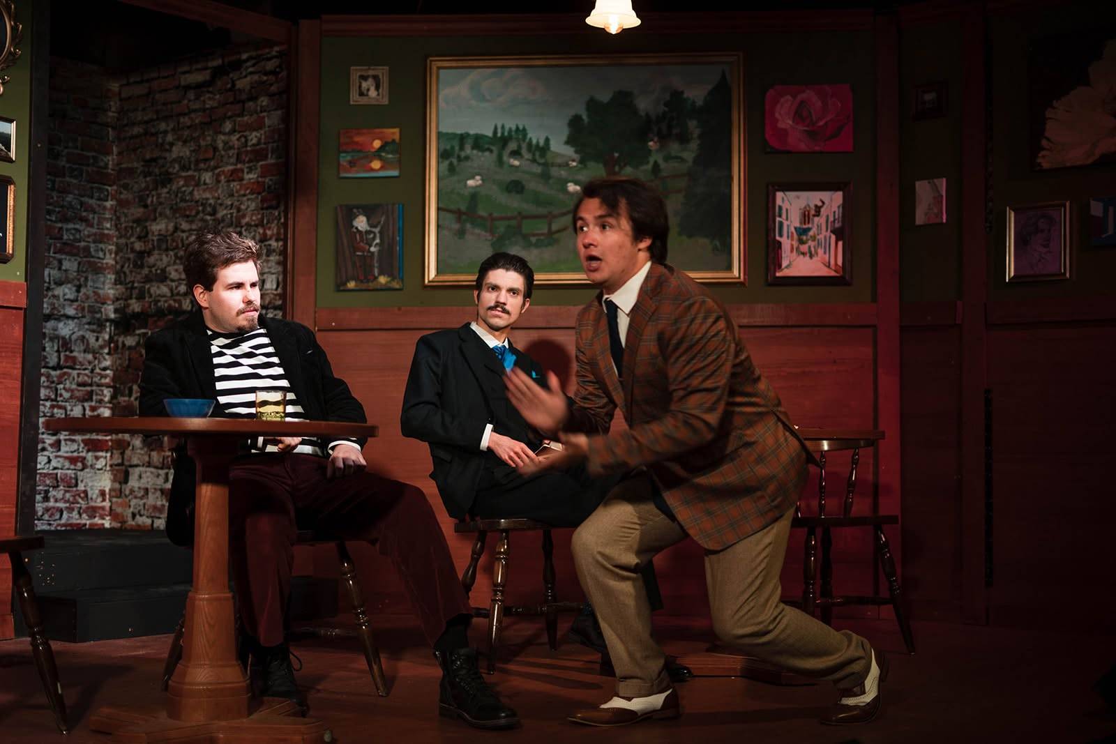 The same performer lunges forward with one hand raised while two other performers sit watching from behind, one in a striped jersey at a table, the other in a dark suit and blue cravat. A large framed landscape hangs on the wall.