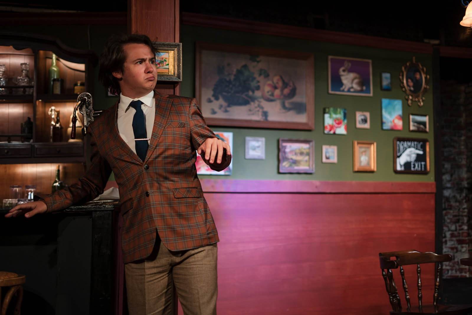 The performer leans against the bar with one hand held out, looking off to the side. The green wall behind carries a framed still life, a wall of small pictures and the “DRAMATIC EXIT” sign.