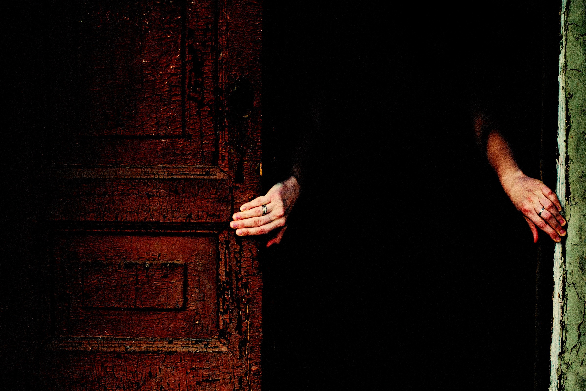 Two hands pushing open a weathered dark red door onto blackness