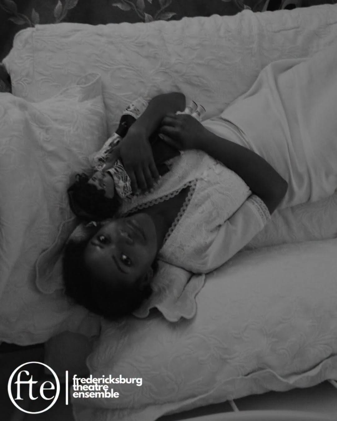 A performer in a white lace nightgown lies on a quilted bed, hugging a doll and looking up at the camera. Black and white photo with the Fredericksburg Theatre Ensemble logo in the lower left.
