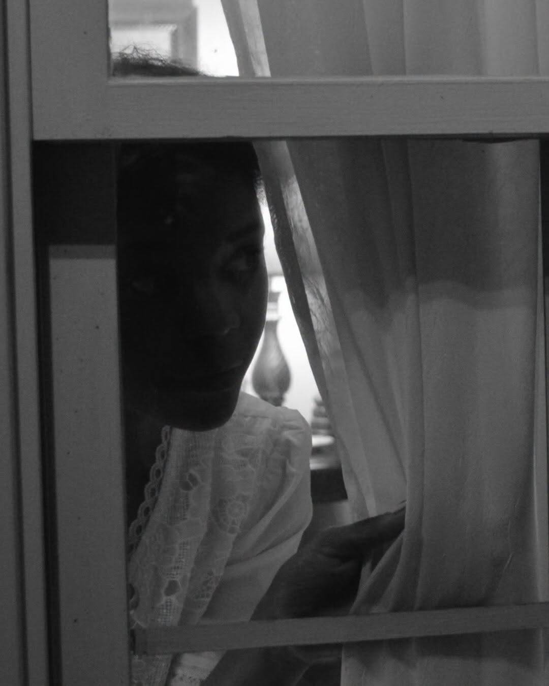 The same performer peers out from behind a sheer curtain through the panes of a window, half in shadow. Black and white photo.