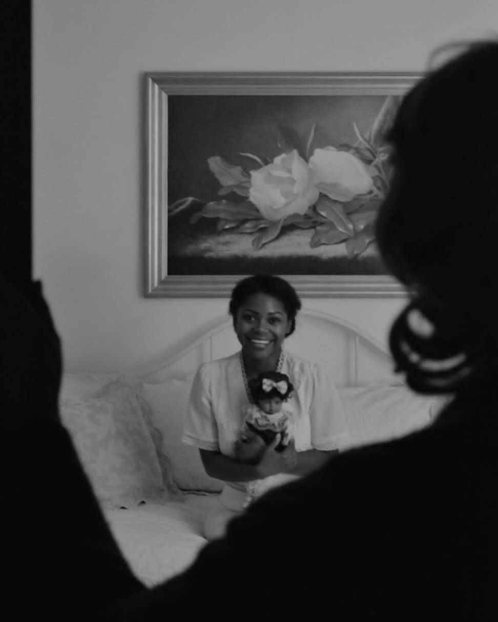 Past the dark silhouette of someone in the foreground, the same performer sits smiling on a white bed holding a doll with a bow, beneath a framed painting of white flowers. Black and white photo.