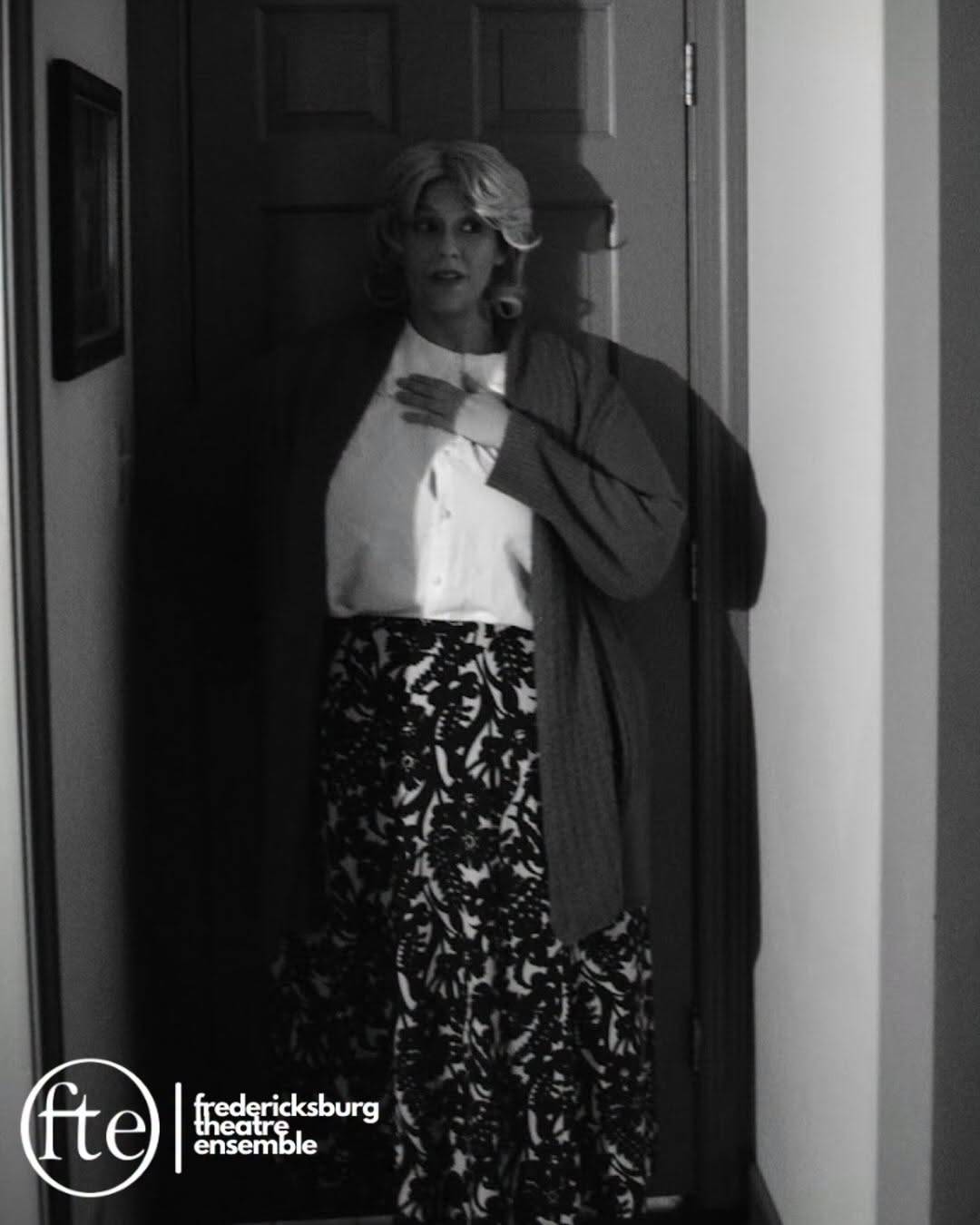 A performer in a blond wig, white blouse, dark cardigan and patterned skirt stands in a doorway with one hand on their chest, glancing to the side. Black and white photo with the Fredericksburg Theatre Ensemble logo in the lower left.