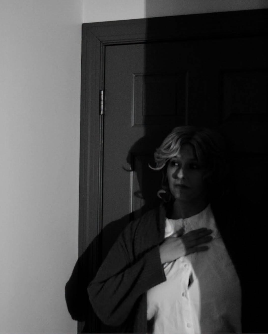 Close view of the same performer in a dim, shadowed doorway, hand on their chest, looking to the side. Black and white photo.