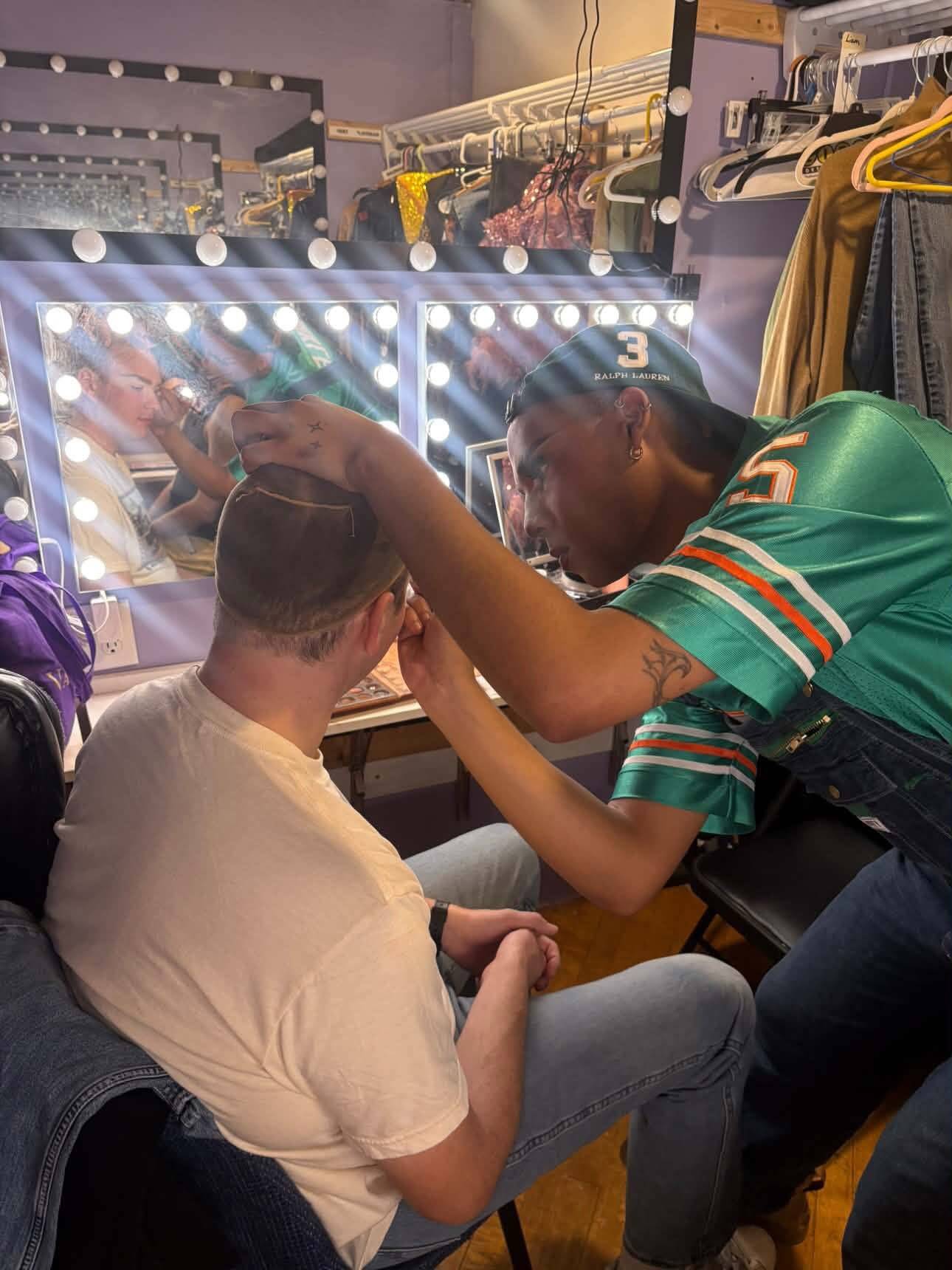 A performer in a green number 5 football jersey and a backwards cap works on the eyebrows of a seated performer in a white T-shirt, in front of a lit makeup mirror and a rack of costumes.