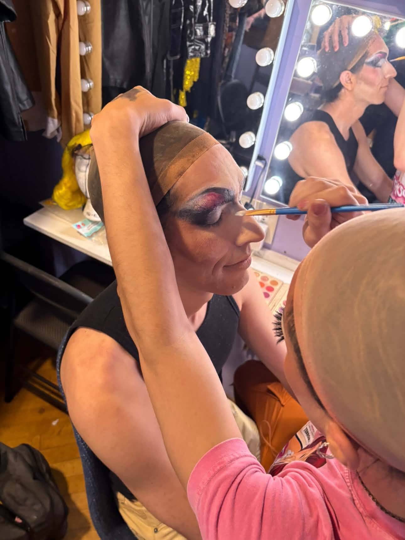 Close view of a seated performer in a wig cap with half-finished purple and pink eye makeup, while a performer in pink paints their brow with a fine brush.