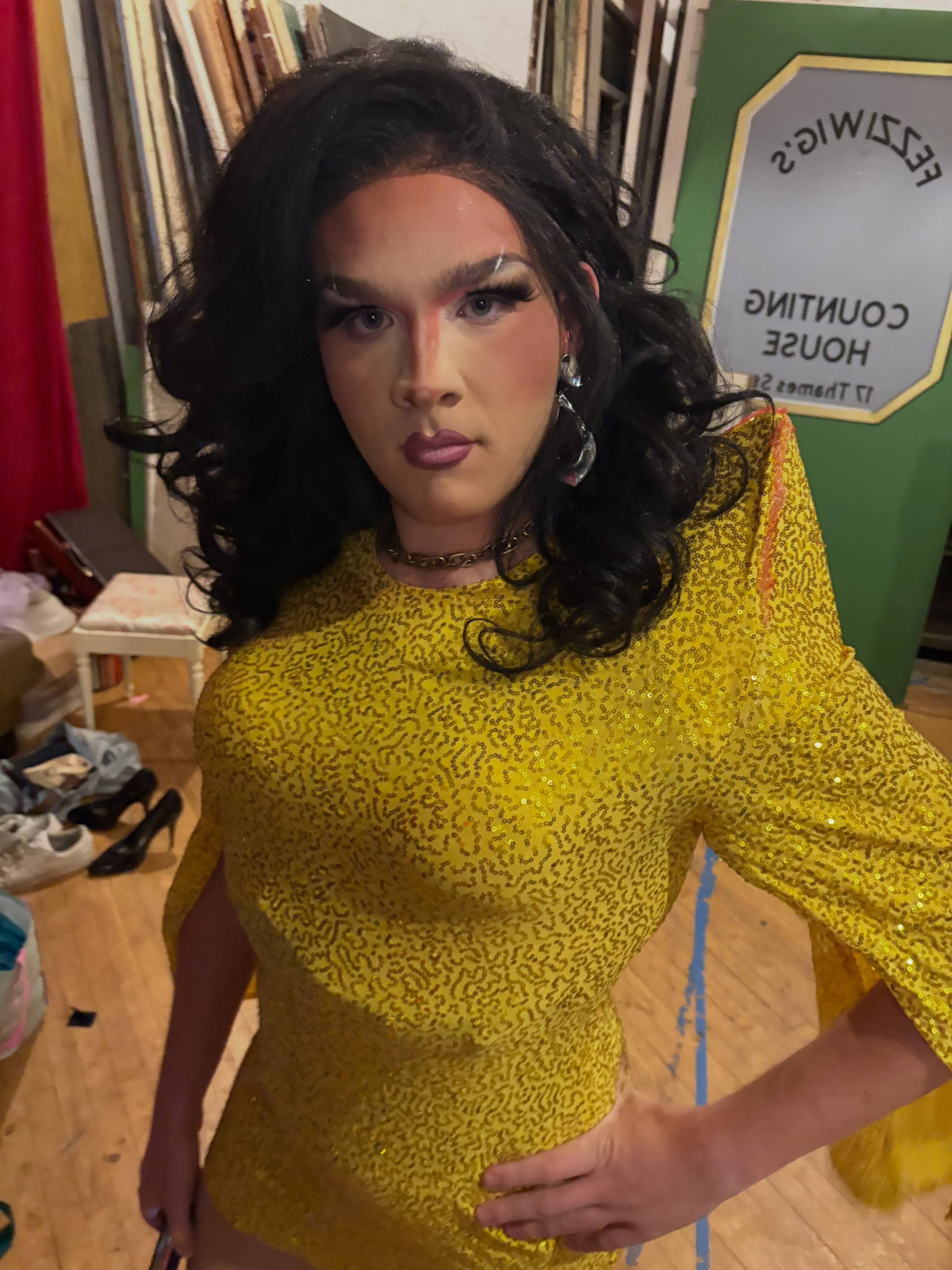 A performer in full drag, in a gold sequined dress with long dark curls and hoop earrings, stands hand on hip backstage beside a painted sign reading Fezziwig’s Counting House.