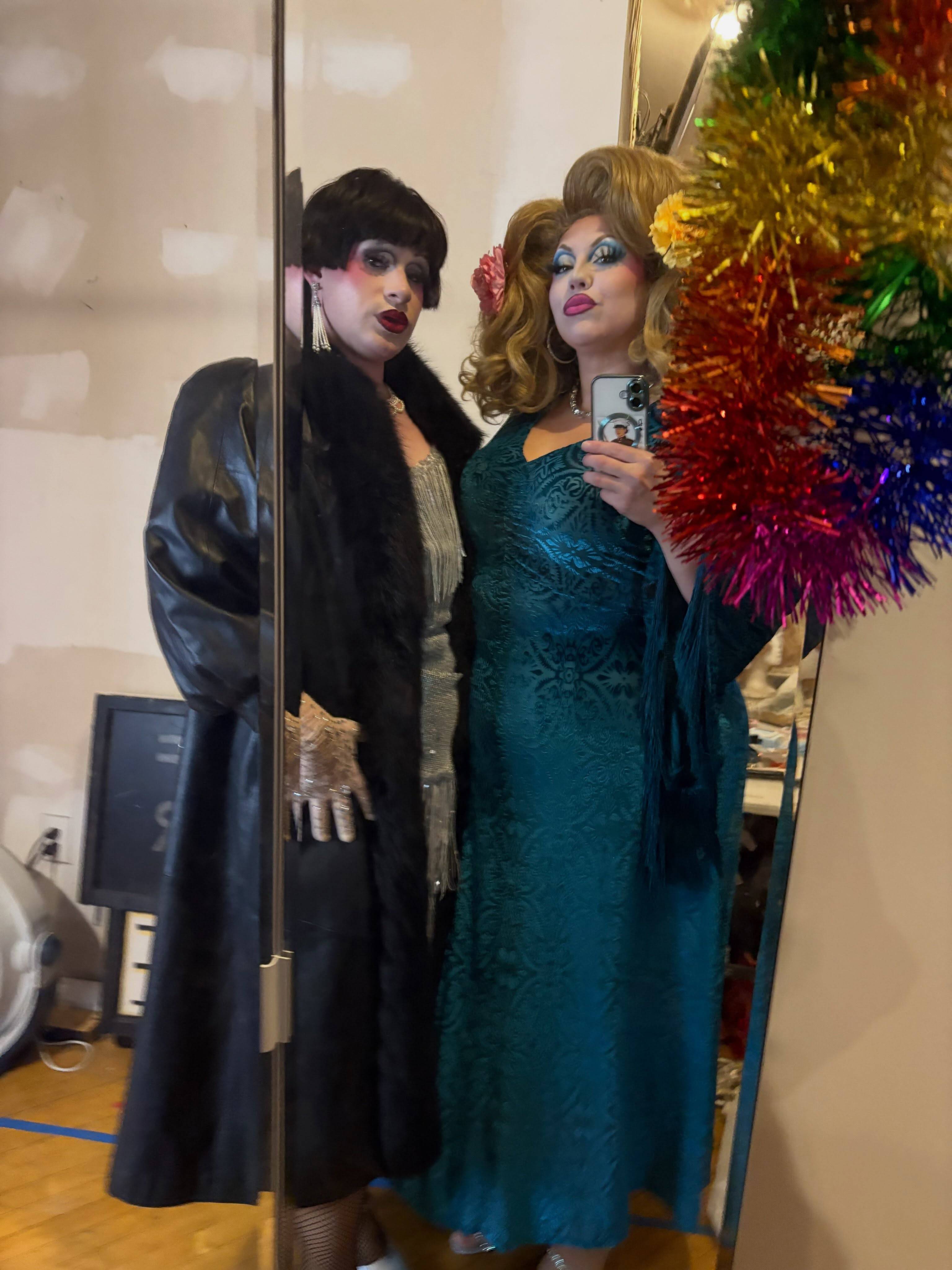 Two performers in drag take a mirror selfie: one in a black leather coat with a fur collar and a dark bob, the other in a teal gown with a blond updo and a flower, beside a rainbow of tinsel.