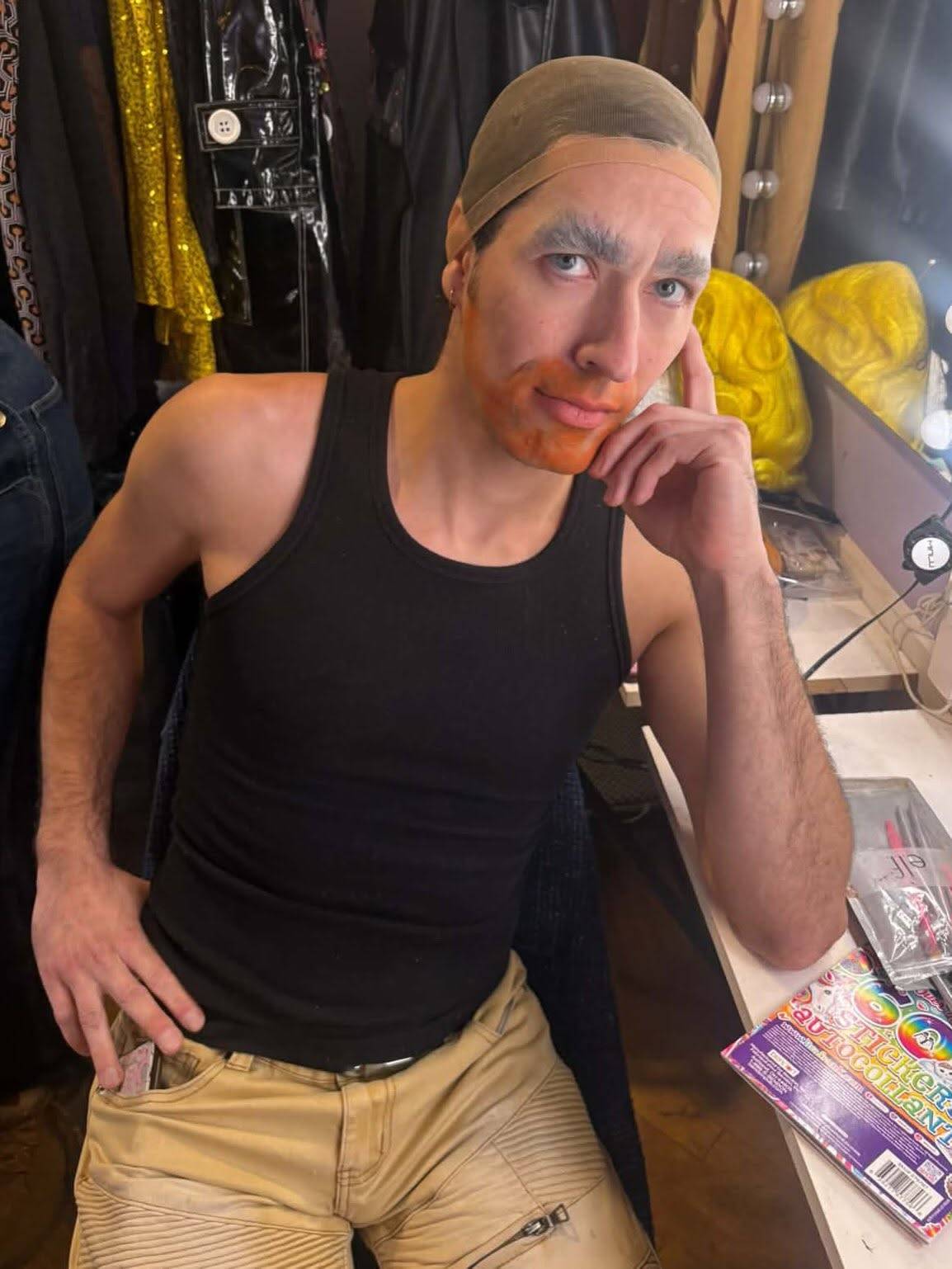 A performer in a wig cap and black tank top, eyebrows blocked out and orange color corrector along the jaw, rests their chin on one hand at a makeup table.
