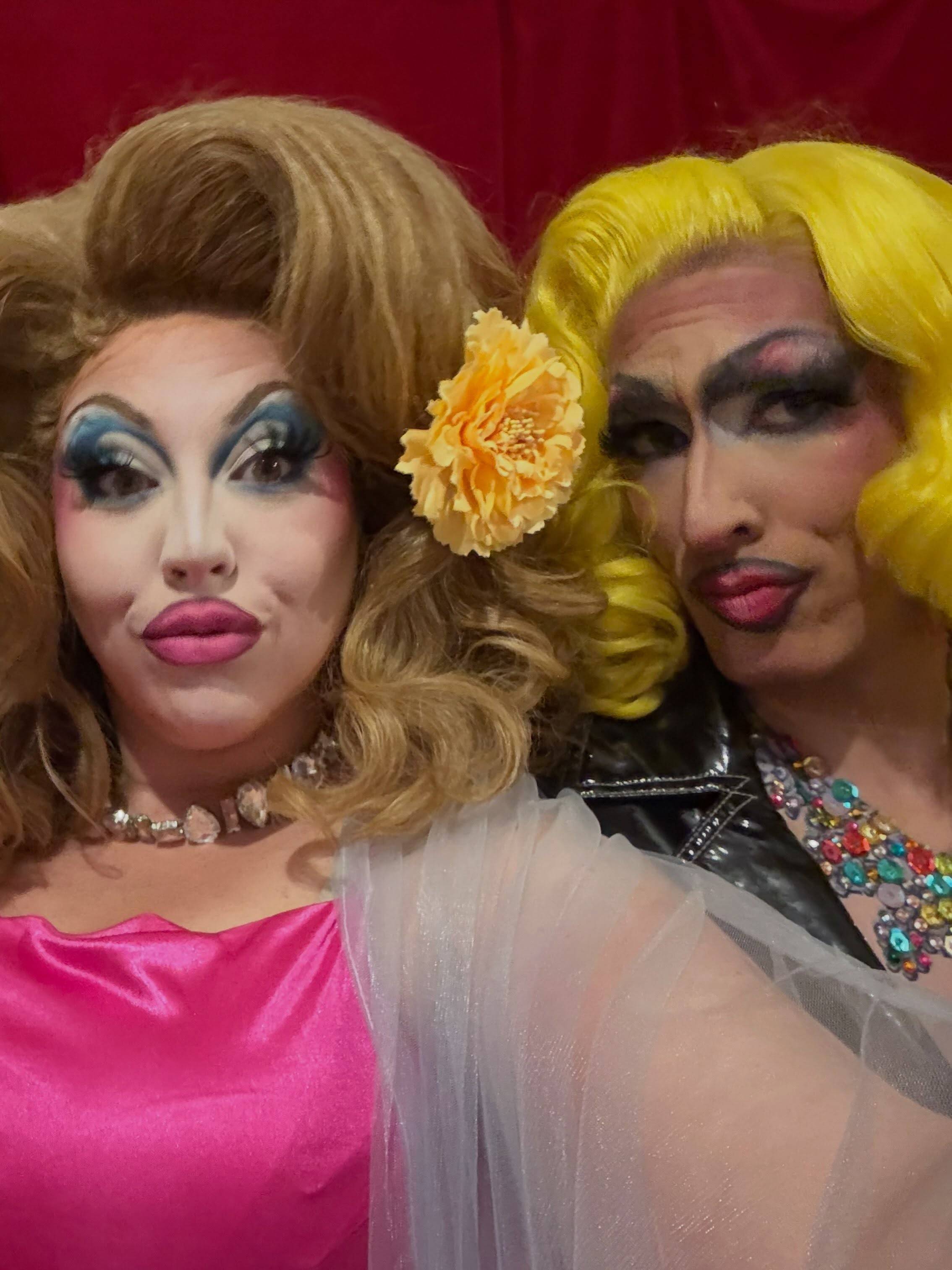 Two performers in full drag pucker up for a selfie against a red curtain: one in pink satin with a honey-blond wig and a yellow flower, the other in a bright yellow wig, leather jacket and jeweled necklace.