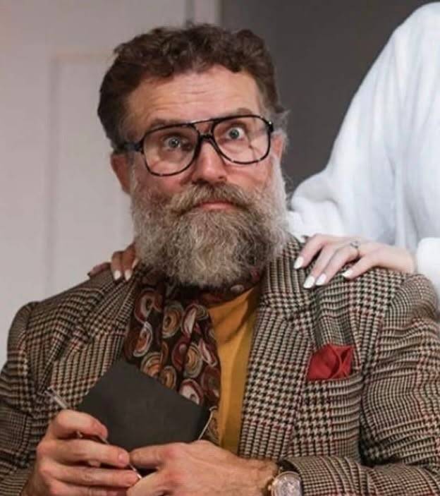 A performer with a full grey beard and tortoiseshell glasses in a tweed jacket, mustard turtleneck, paisley cravat and red pocket square, holding a small notebook and pen; another person’s hand rests on their shoulder.